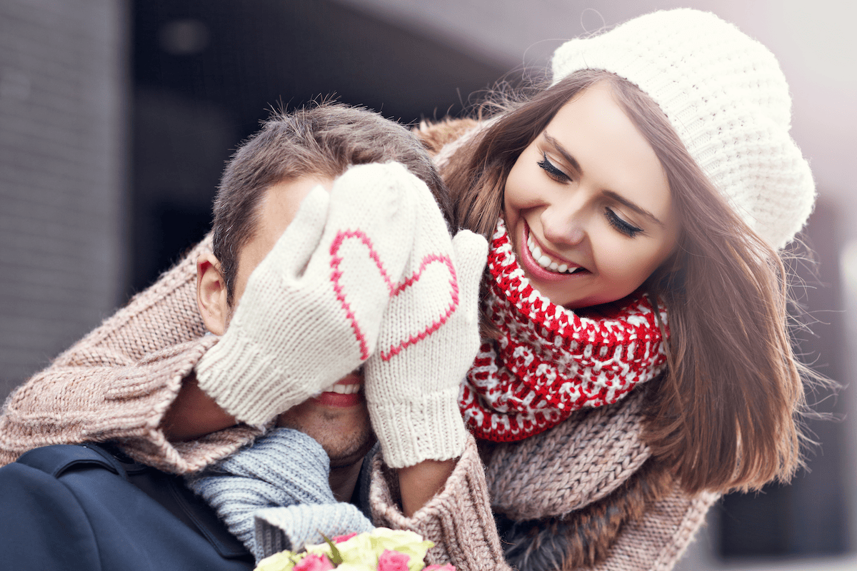 Young woman holding mittens over her boyfriends eyes that have a heart on them