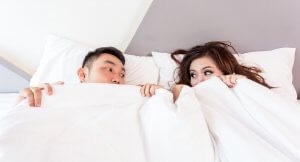 Couple whose heads are sticking up out of bed covers look at each other.