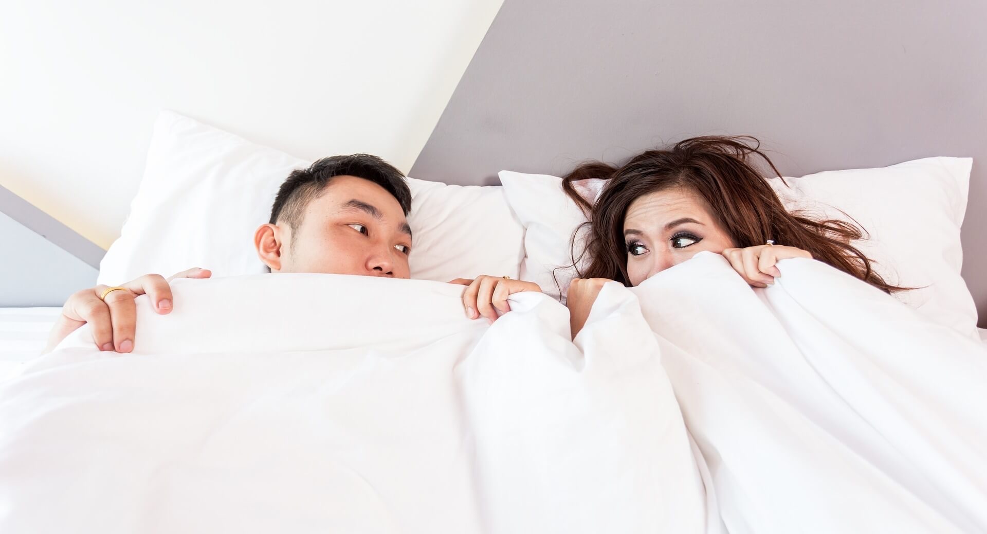 Couple whose heads are sticking up out of bed covers look at each other.