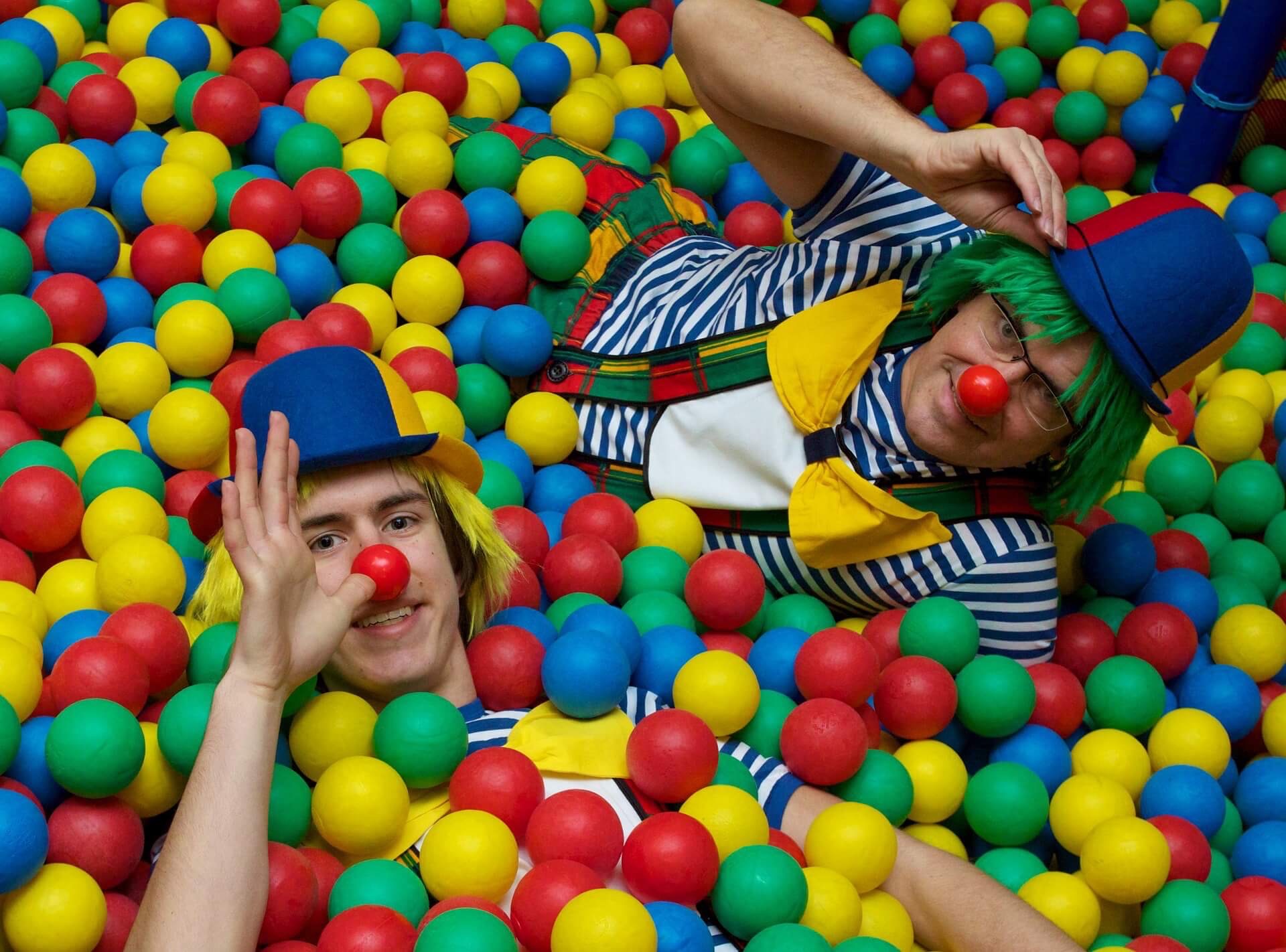 Two clowns immersed in colorful plastic balls, one clown thumbing his nose. Photo illustration