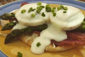 Two oven-‘poached‘ eggs nestled on top of a toasted multi-grain flatbread with melted Havarti cheese, oven-crisped bacon, asparagus spears, topped off with a béchamel sauce, and a few scallions for garnish.