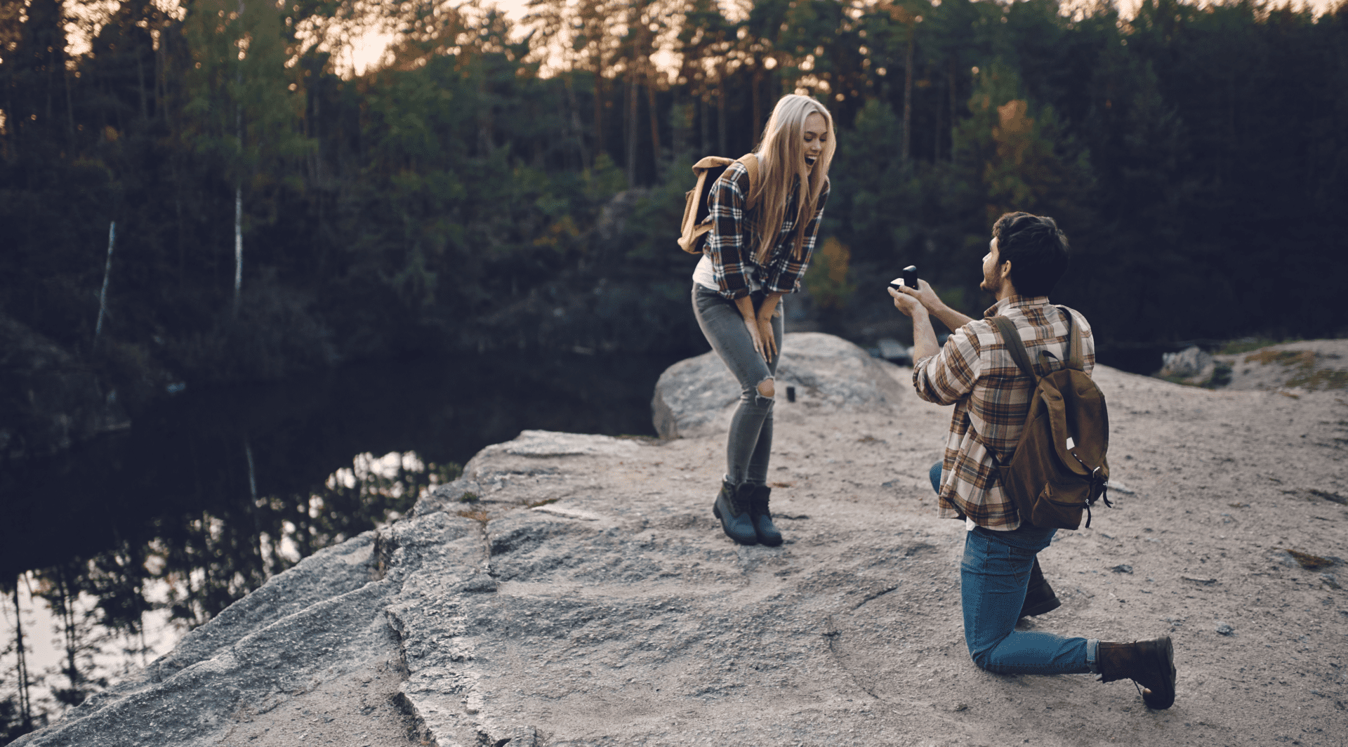 Young male adult on his knees proposing to a woman in a plaid shirt beside a lake